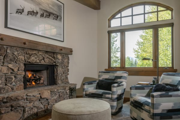 Den with a stone fireplace, two patterned armchairs and a round ottoman by an arched window