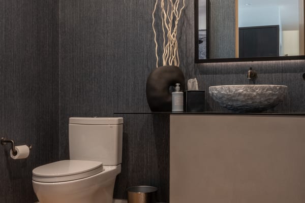 Powder room with dark textured wallpaper, a stone vessel sink and a backlit mirror