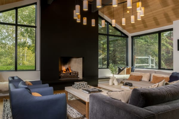 Living room with a lit fireplace, ring chandelier, wood ceiling and tall windows onto the garden