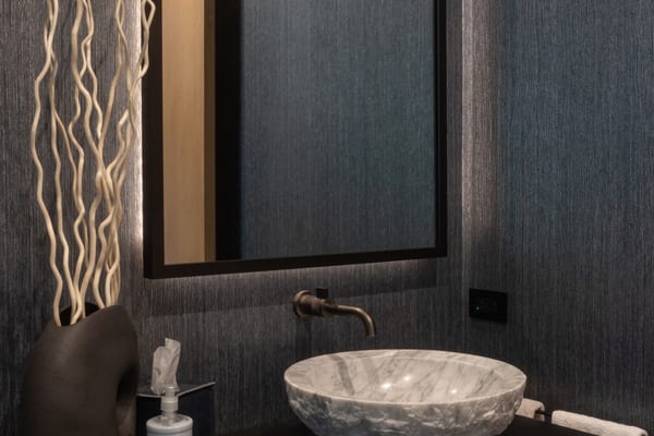 Stone vessel sink and bronze wall faucet below a backlit mirror
