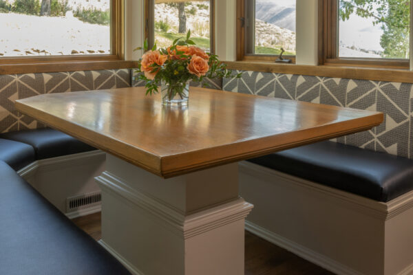 Breakfast nook with wrap-around booth seating. A bouquet of peach roses is displayed on the table. The nook is surrounded on all three sides by large windows with a view of the trees and mountains.