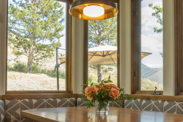 Breakfast nook with wrap-around booth seating. A bouquet of peach roses is displayed on the table. Various log beams meet to sustain a hanging lamp. The nook is surrounded on all three sides by large windows with a view of the trees and mountains.