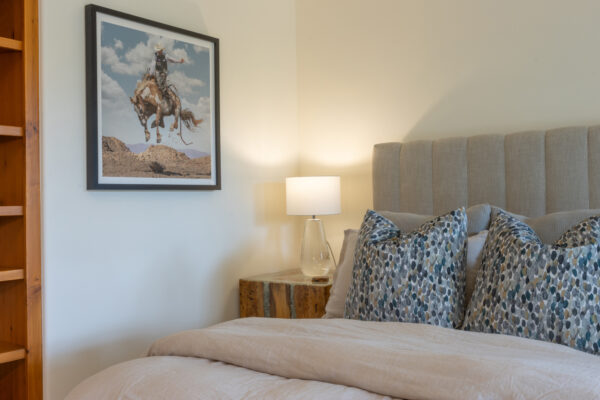 Close up of bed with decorative pillows, side table with lamp, artwork of a bucking horse with rider hangs on the wall, and built-in wall shelves.